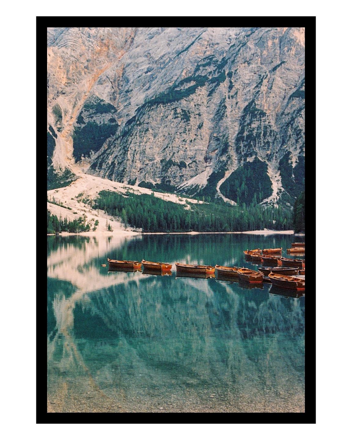 Wooden boats are moored on a clear lake with mountains and a forest in the background.