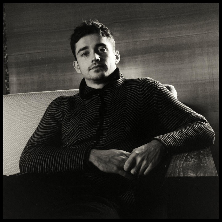 A man with short hair and a patterned sweater sits on a couch with his hands clasped.