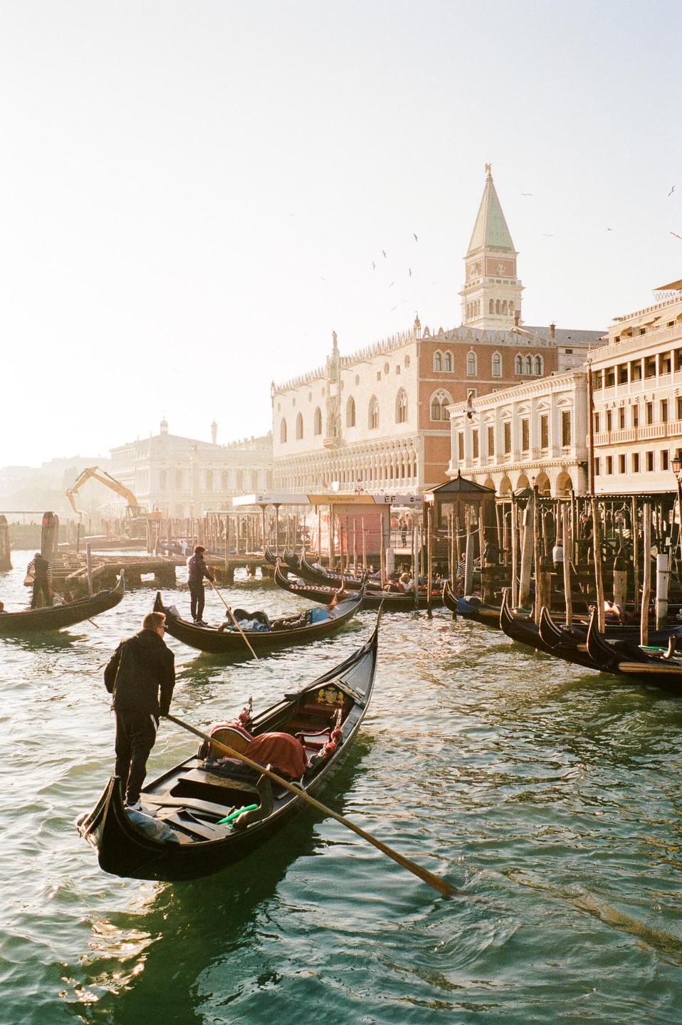 Gondoliers navigate their boats through a canal in Venice, with historic buildings and a bell tower visible in the background.