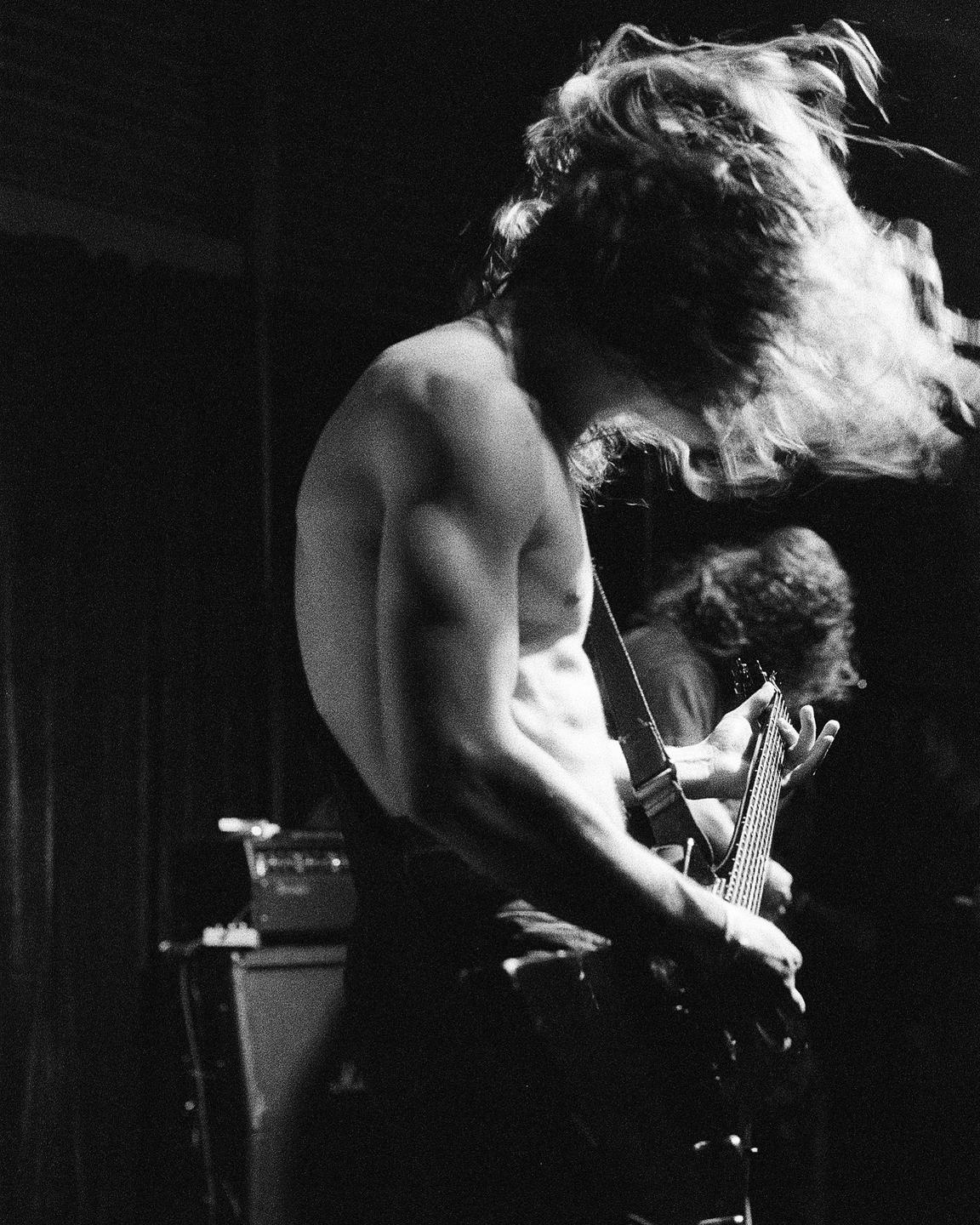 A shirtless man with long hair plays an electric guitar on a dark stage.
