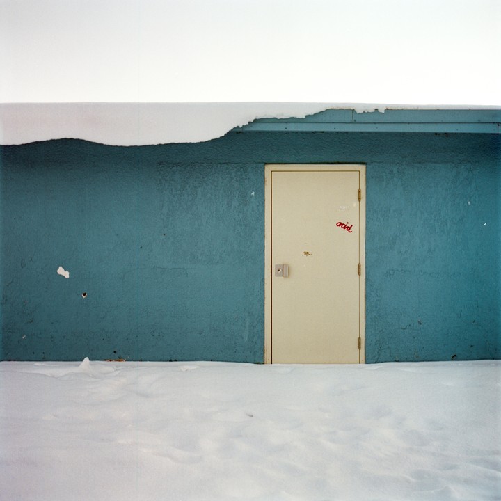 A cream-colored door with the word "acid" written on it is set into a blue wall, with snow covering the ground and the top of the wall.
