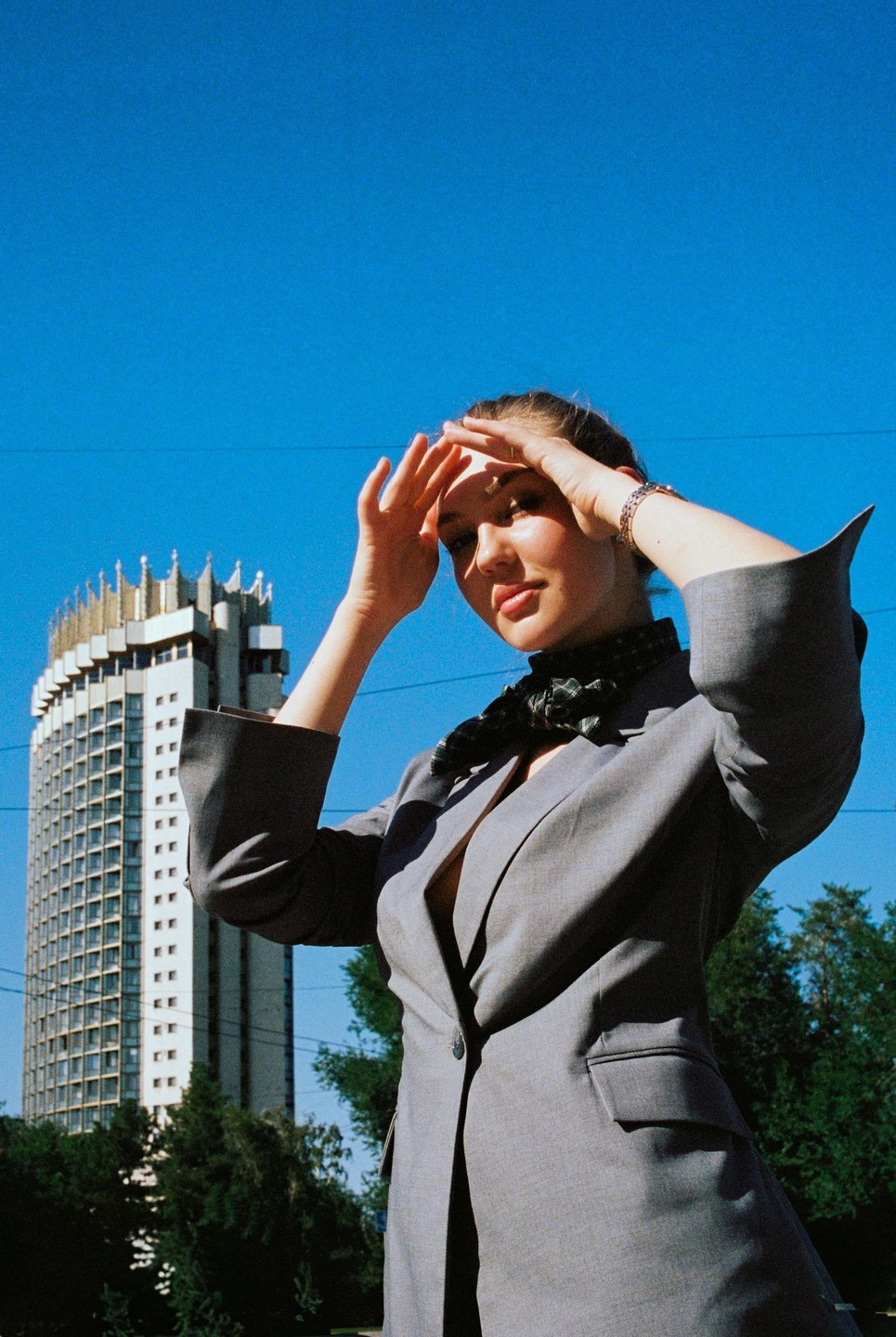 A woman shields her eyes from the sun in front of a tall building with a decorative roof.