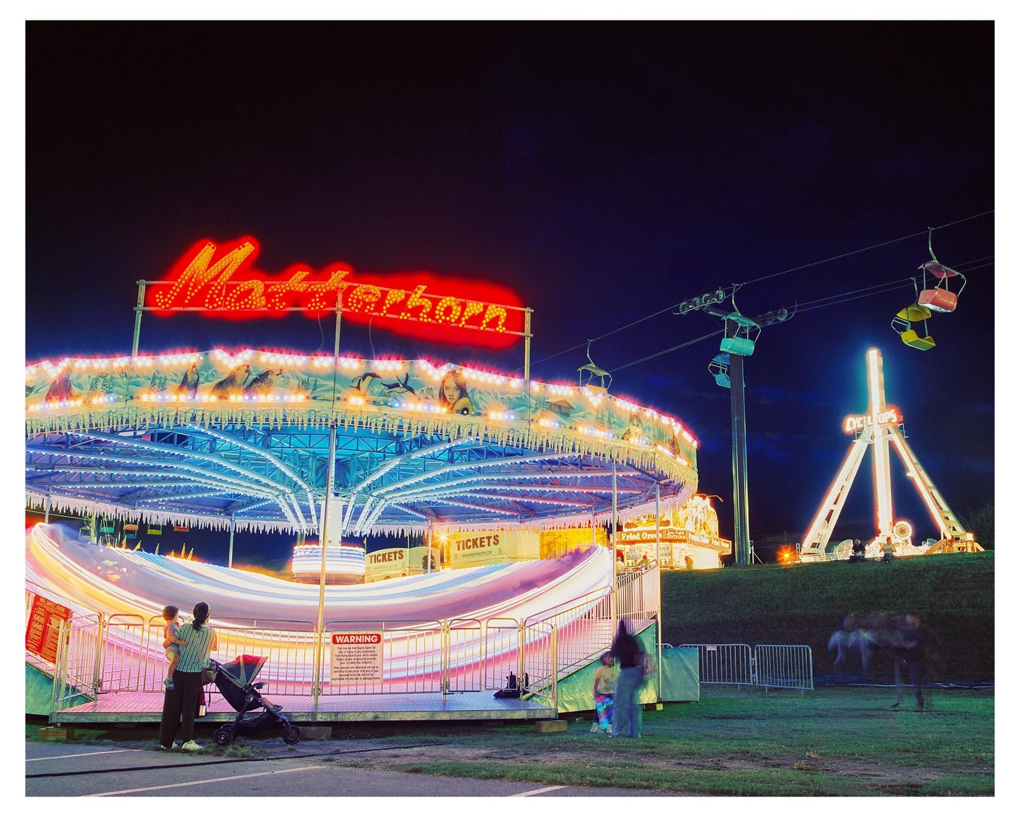 A fairground at night features a spinning Matterhorn ride with blurred lights, a ski lift, and a Cyclops ride in the background, with people watching the rides.