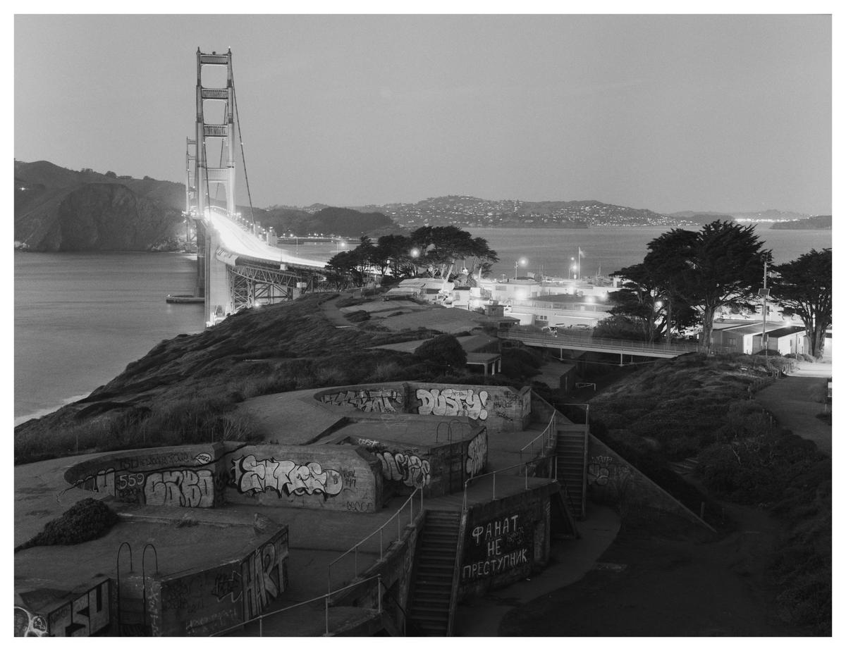 A black and white landscape shows the Golden Gate Bridge at night, with graffiti-covered bunkers in the foreground and a city across the water.