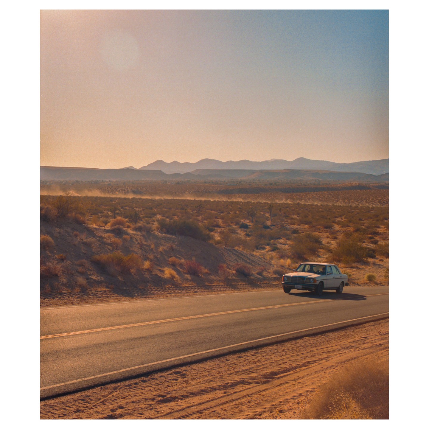 A white car drives on a desert road with mountains in the distance.