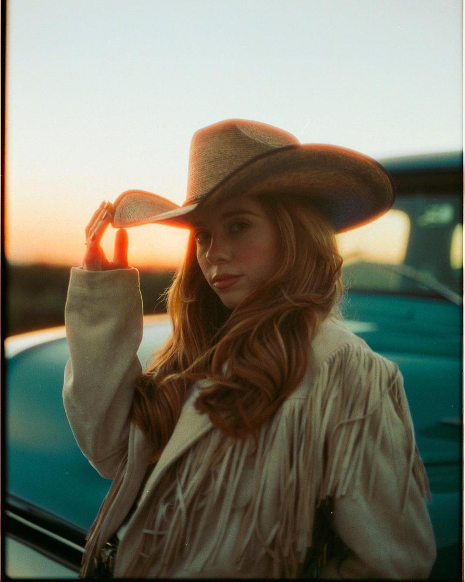 A woman wearing a cowboy hat and a fringed jacket stands in front of a car at sunset.