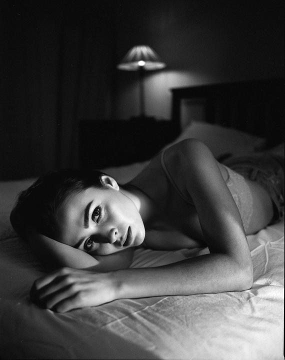 A woman lies on a bed with her head resting on her arm, looking towards the viewer.
