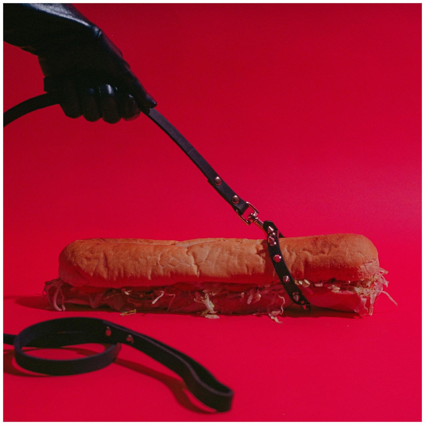 A gloved hand holds a leash attached to a sub sandwich on a red background.