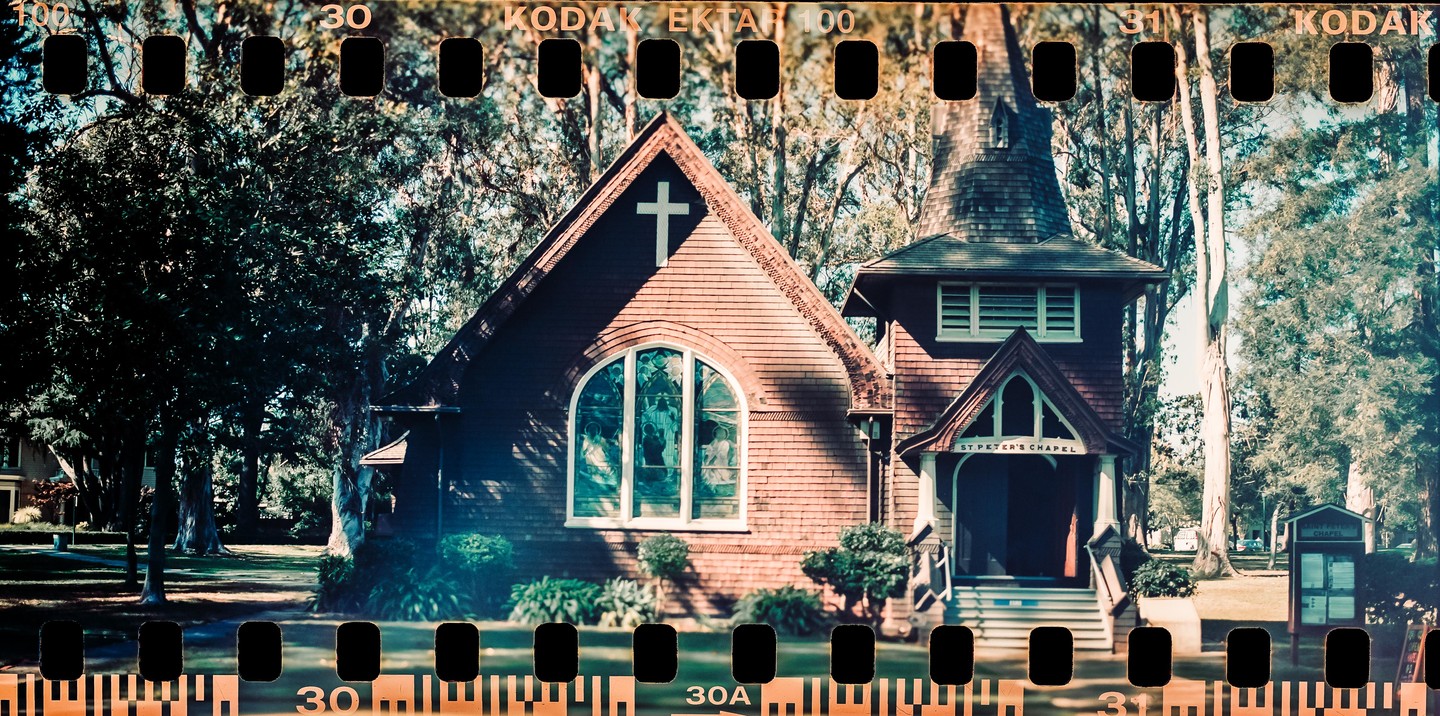 A small chapel with a cross on its facade and a stained glass window is surrounded by tall trees.