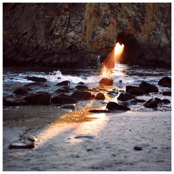 Sunlight streams through a rock arch onto a beach with waves and rocks.