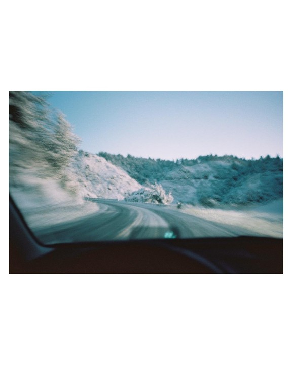 A winding road is seen from inside a vehicle, with snow-covered hills and trees on either side under a clear sky.