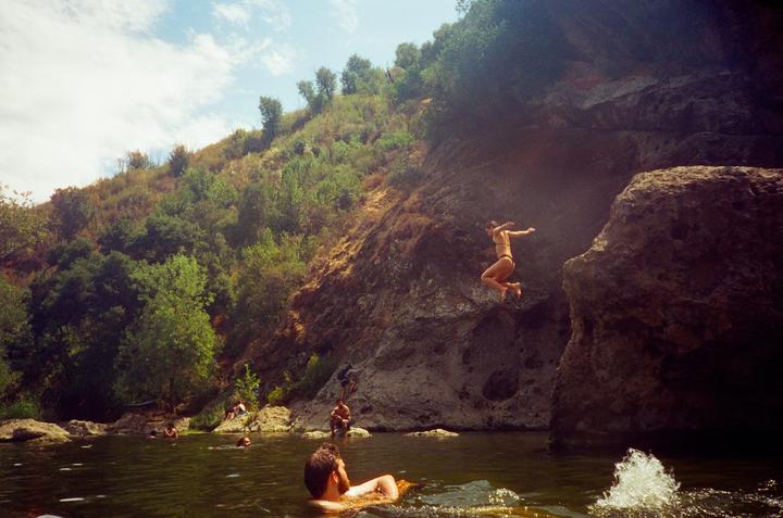 A woman jumps from a rock into a swimming hole with other people in the water and on the shore.