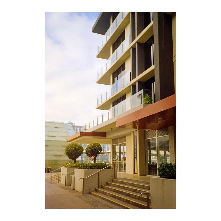 A modern building with balconies and glass railings stands next to a set of stairs leading to its entrance, with two manicured trees in planters.