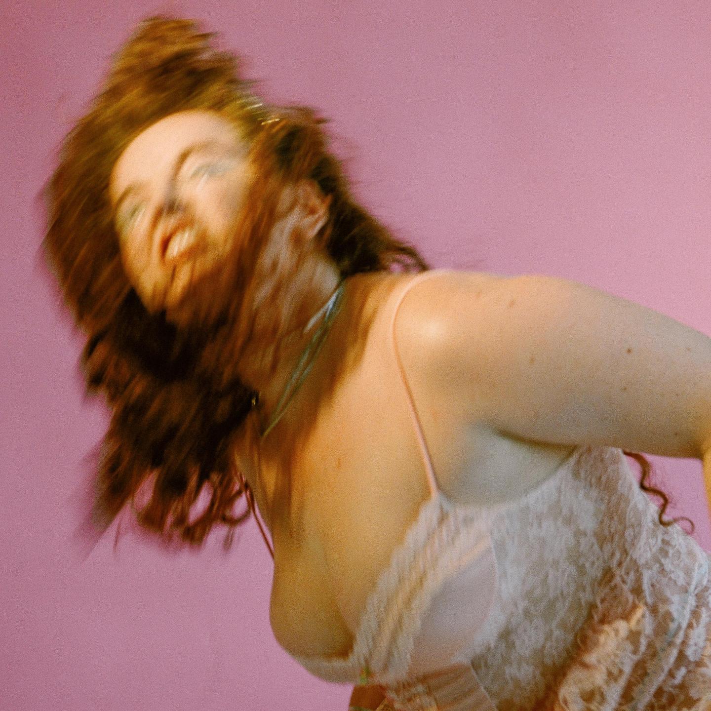 A woman with long hair and a lace top is captured in motion against a pink background.
