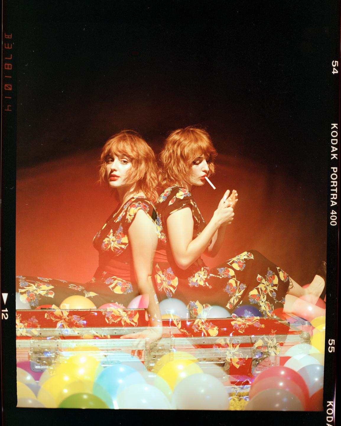 A woman with red hair is sitting back to back with her double exposure, who is lighting a cigarette, surrounded by colorful balloons.