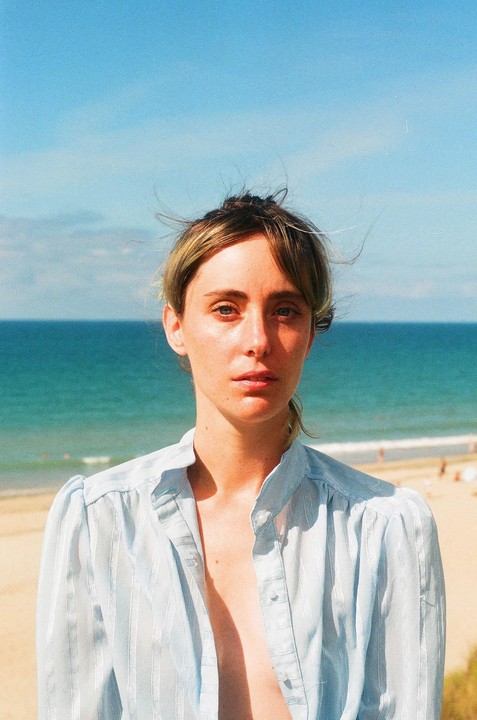 A woman with her hair tied up and wearing an unbuttoned shirt stands on a beach with the ocean behind her.