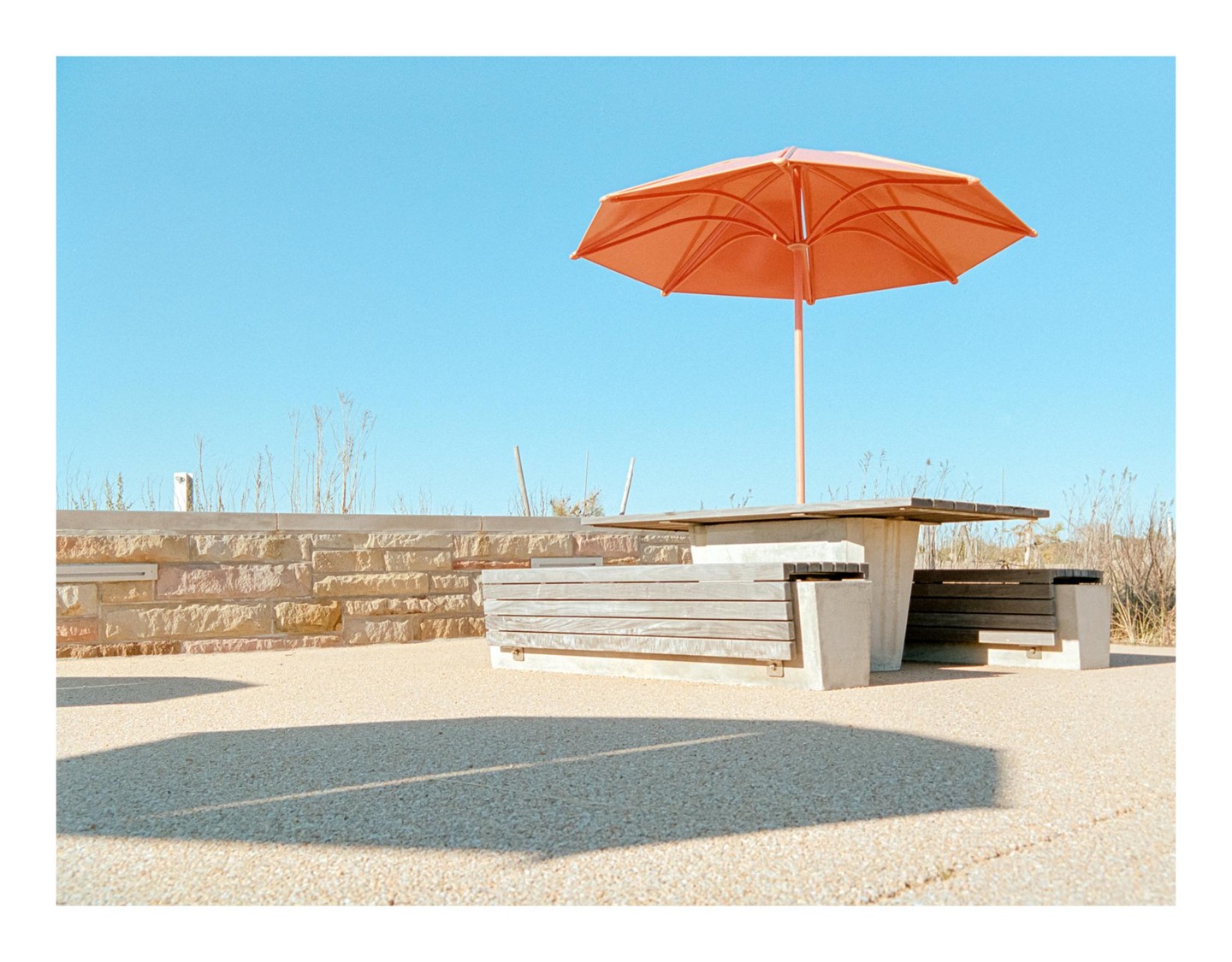 An orange umbrella shades a picnic table and benches next to a stone wall under a clear blue sky.