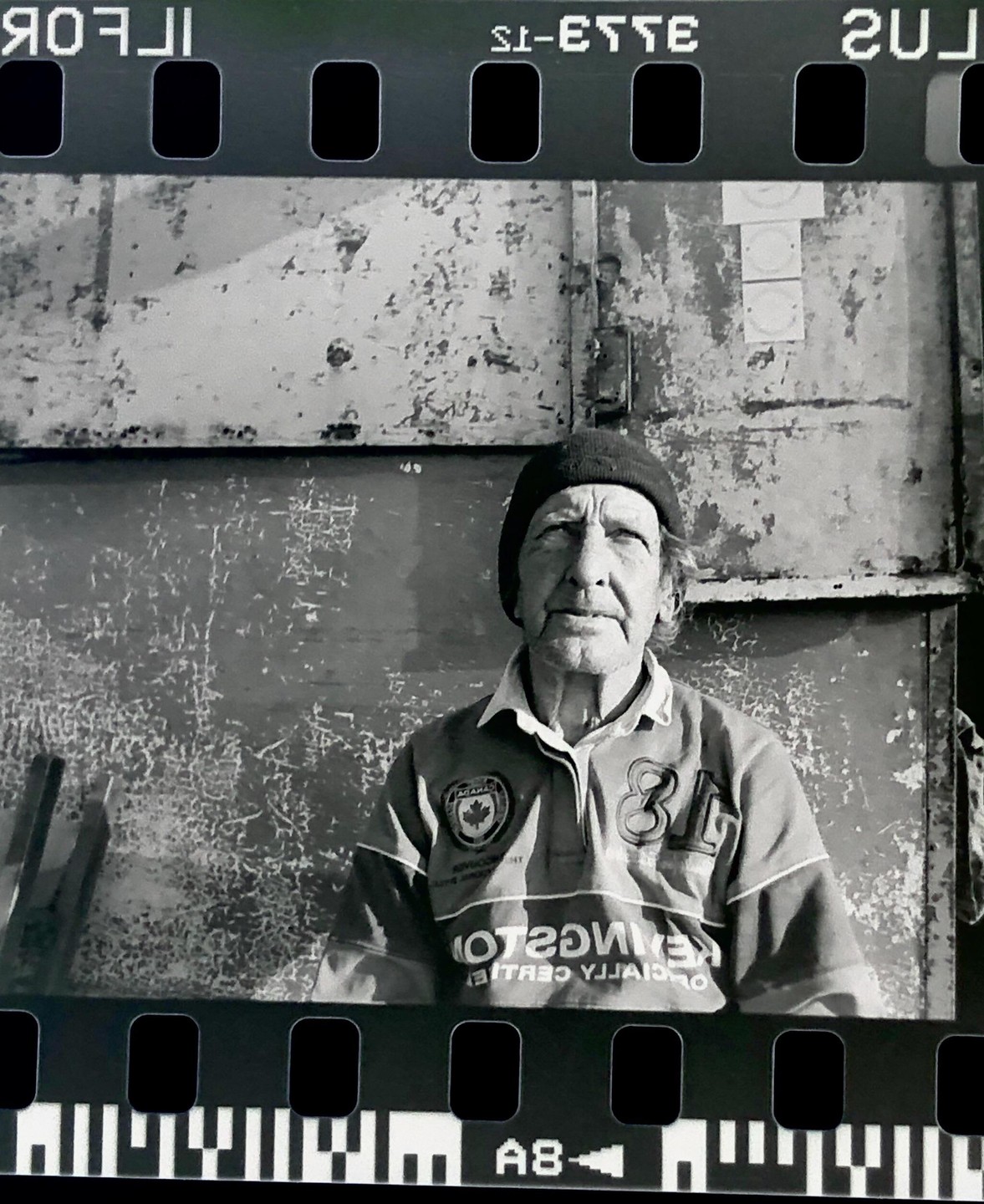 A man wearing a beanie and a rugby shirt is sitting in front of a distressed metal wall.