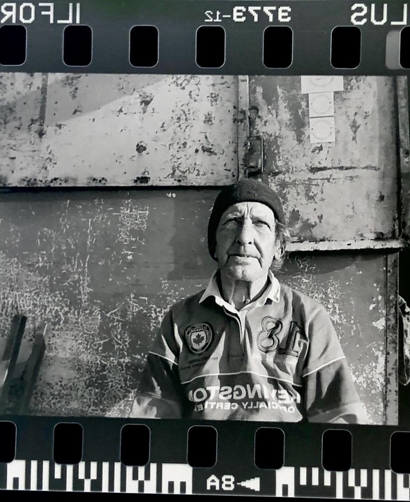 A man wearing a beanie and a rugby shirt is sitting in front of a distressed metal wall.