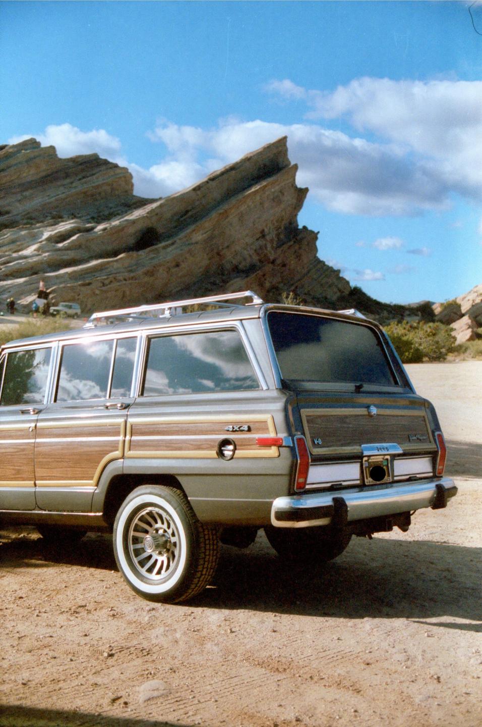 A gray station wagon with wood paneling is parked on a dirt road in front of a large rock formation under a blue sky with clouds.