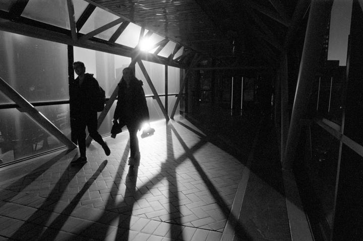 Two people walk through a modern glass and metal walkway with strong backlighting and shadows.