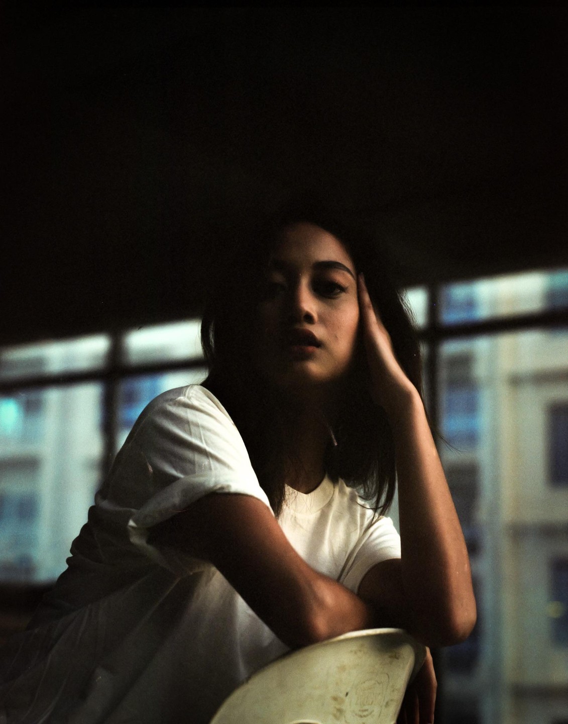 A woman in a white shirt sits with her arm resting on a chair, looking towards the viewer.