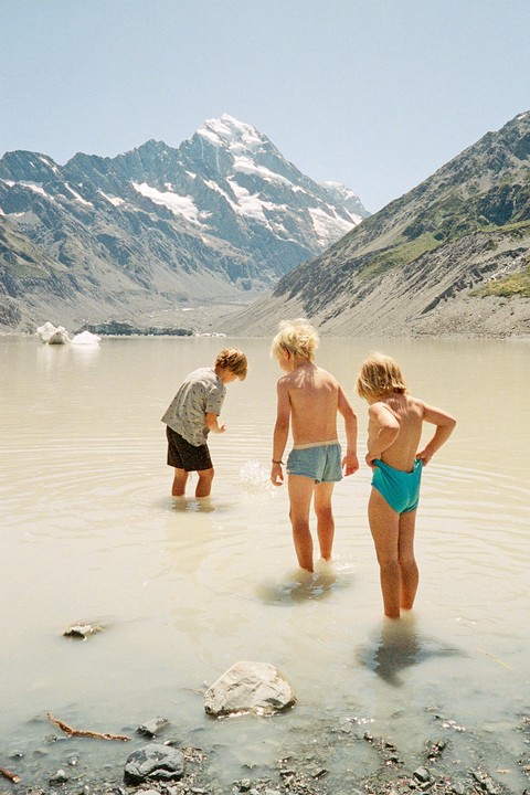 Three children stand in a shallow lake with snow-capped mountains in the background.