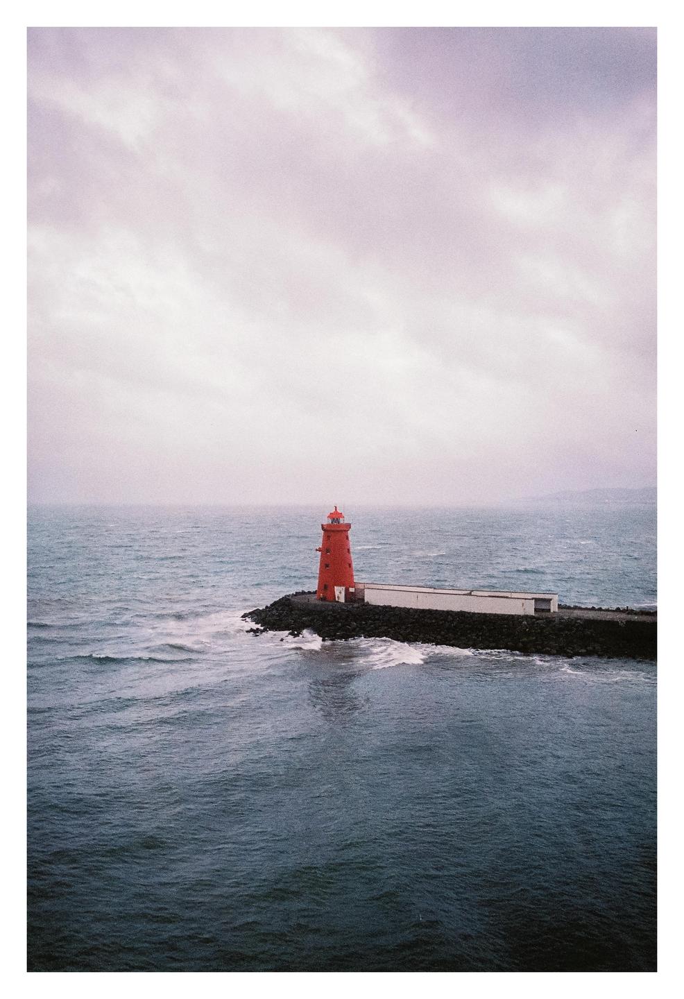 A red lighthouse stands on a rocky pier next to a white building, with waves crashing against the rocks and a cloudy sky above.