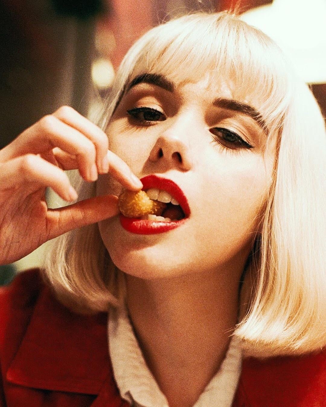 A woman with red lipstick and blonde hair is putting a small, round, fried food item into her mouth.