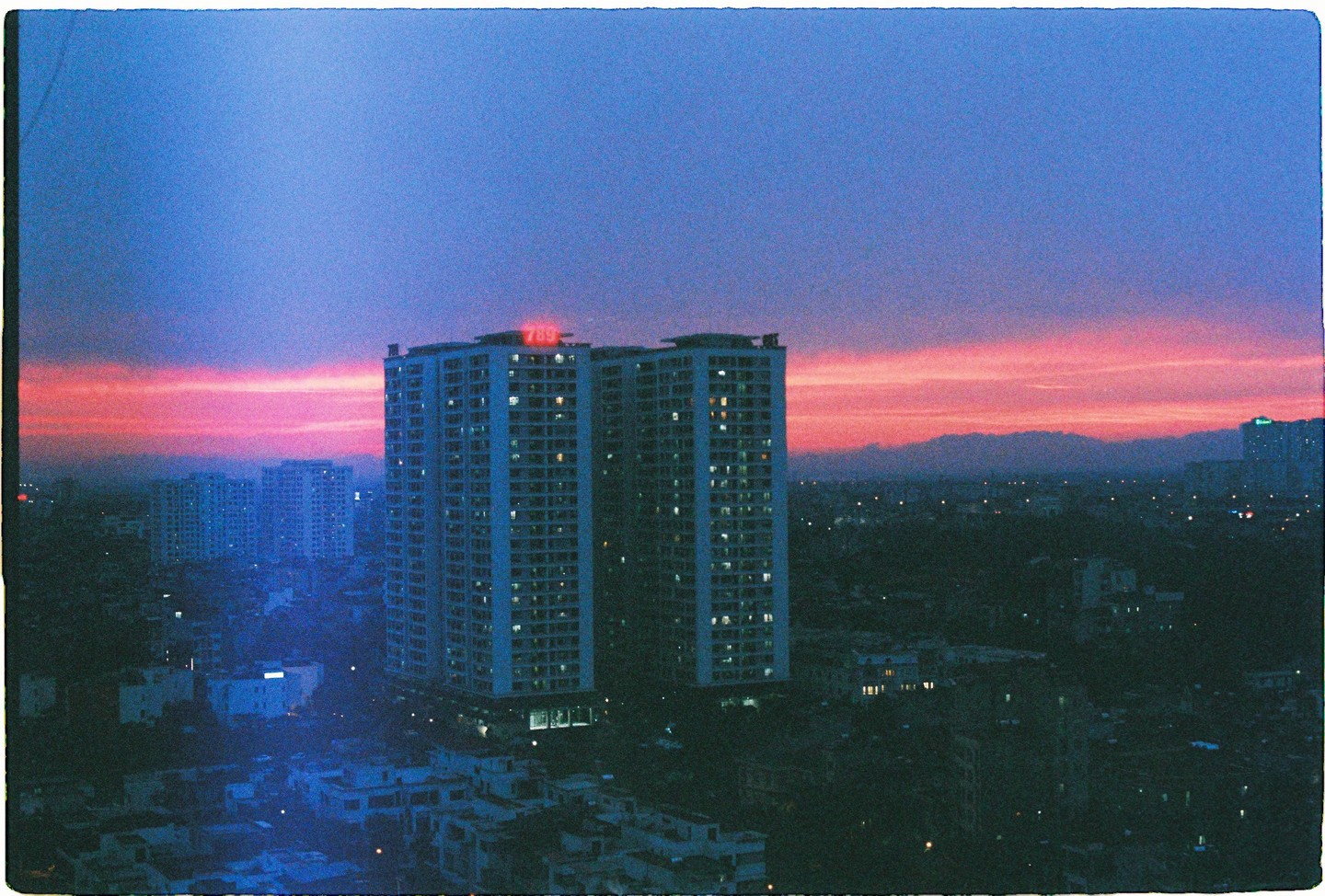 A cityscape at dusk shows tall buildings with illuminated windows against a sky with a vibrant pink and purple sunset.
