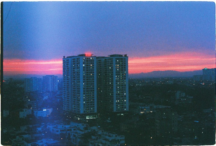 A cityscape at dusk shows tall buildings with illuminated windows against a sky with a vibrant pink and purple sunset.