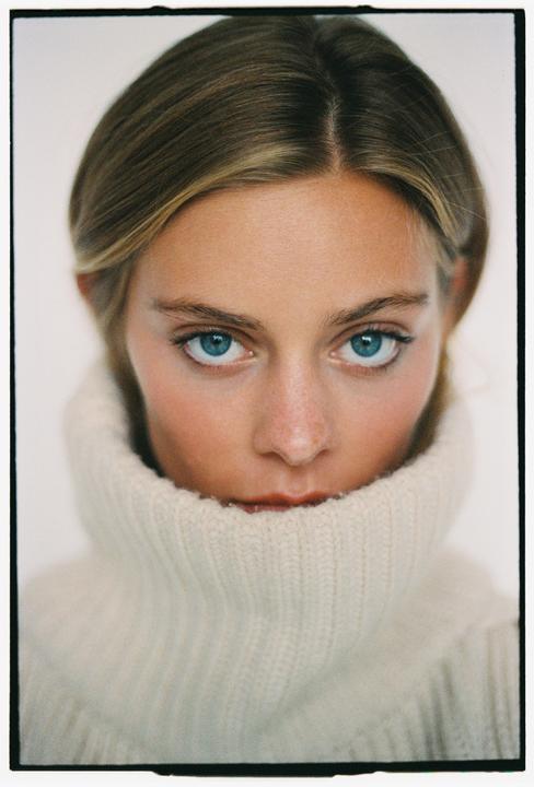 A woman with blue eyes looks directly at the viewer, her face partially obscured by a cream turtleneck sweater.