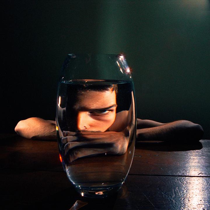 A man's face is distorted through a glass of water on a wooden table.