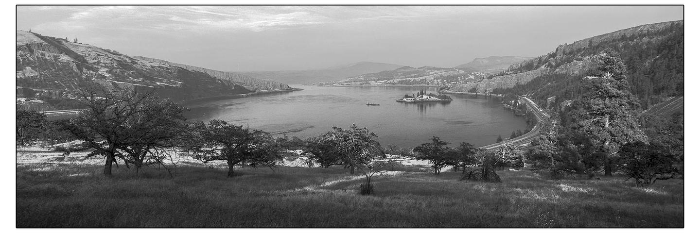 A panoramic black and white landscape shows a wide river with a small island featuring buildings, surrounded by mountains and a road following the riverbank.