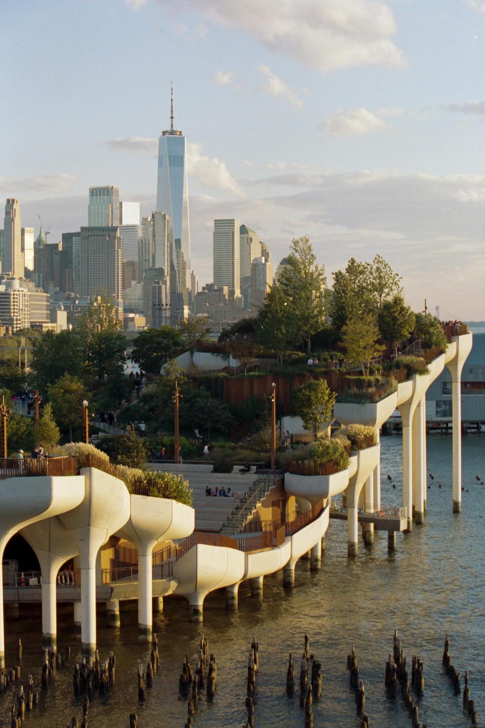 A modern park with trees and people is built on stilts over water, with a city skyline in the background.