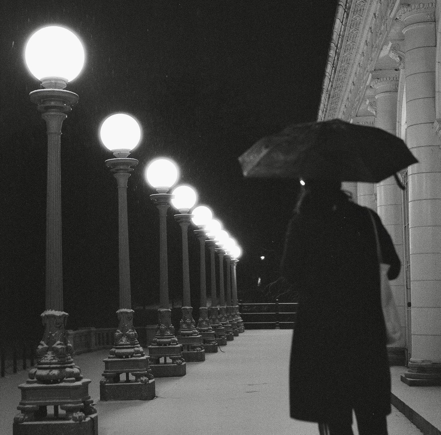 A person with an umbrella walks past a row of glowing streetlights on a snowy night.