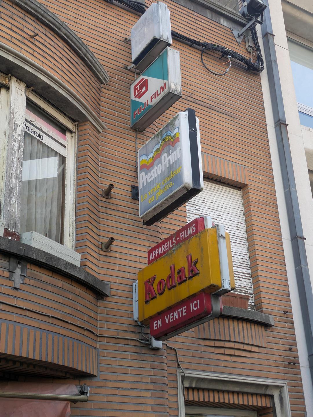 Several old advertising signs for film and photo processing are mounted on a brick building.