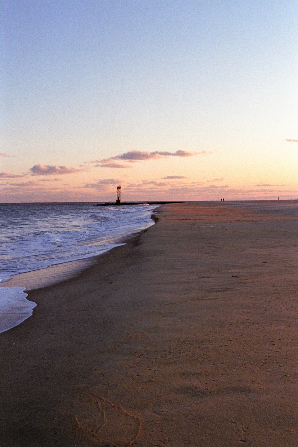 A beach with a lighthouse in the distance is shown at sunset.
