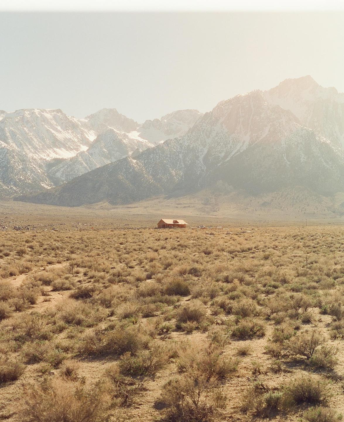 A wooden house sits in a vast, arid landscape with snow-capped mountains in the background.