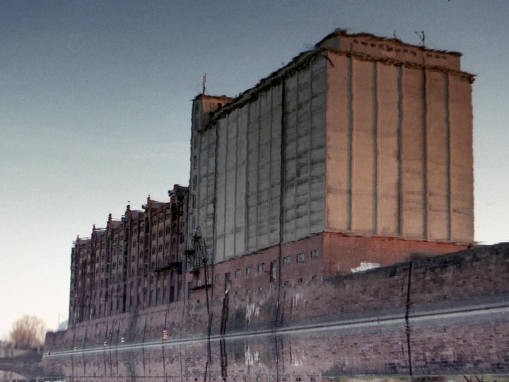A large industrial building and a row of smaller buildings are reflected in water under a pale sky.
