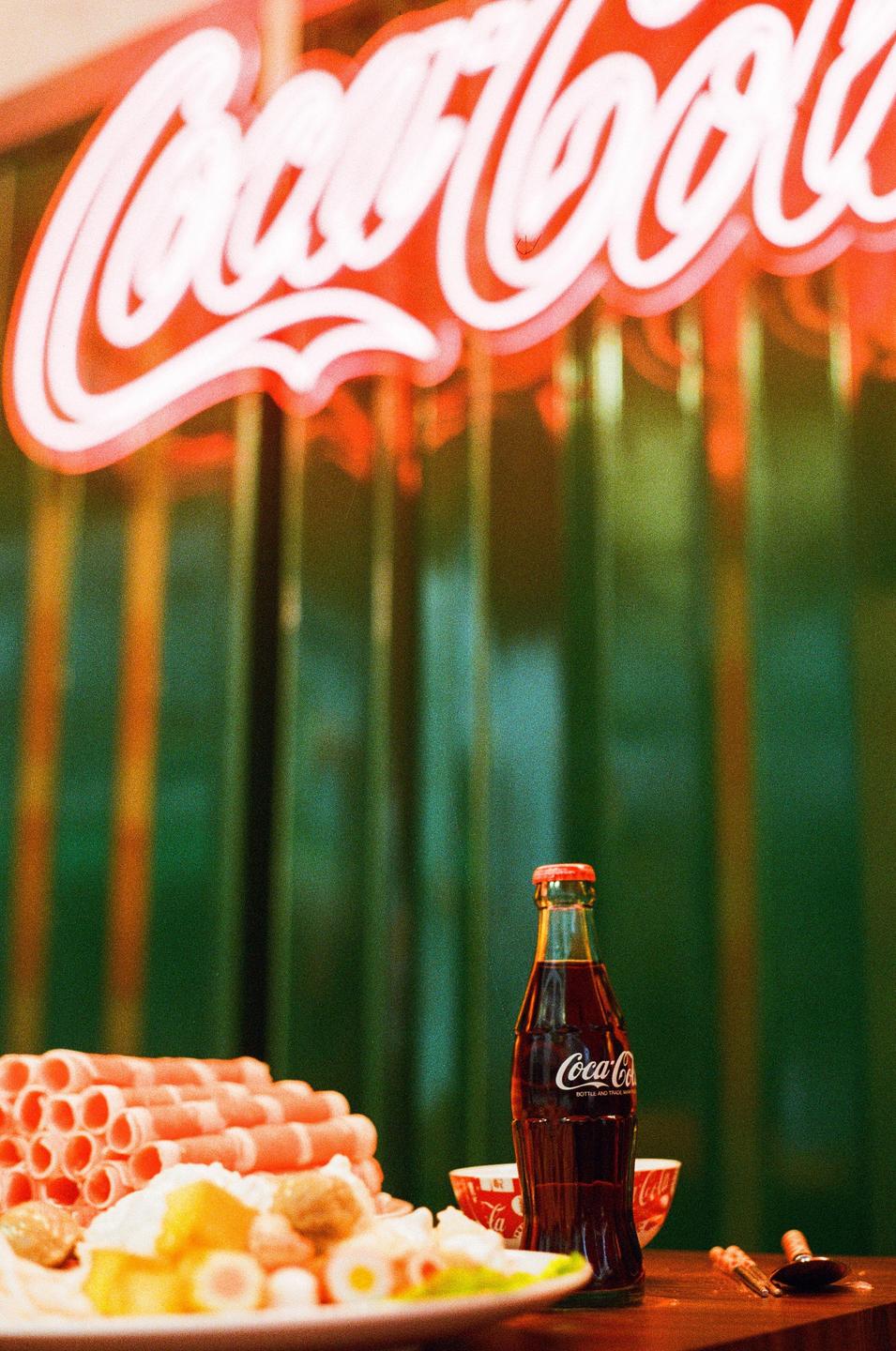 A Coca-Cola bottle sits on a table next to a plate of food with a neon Coca-Cola sign in the background.