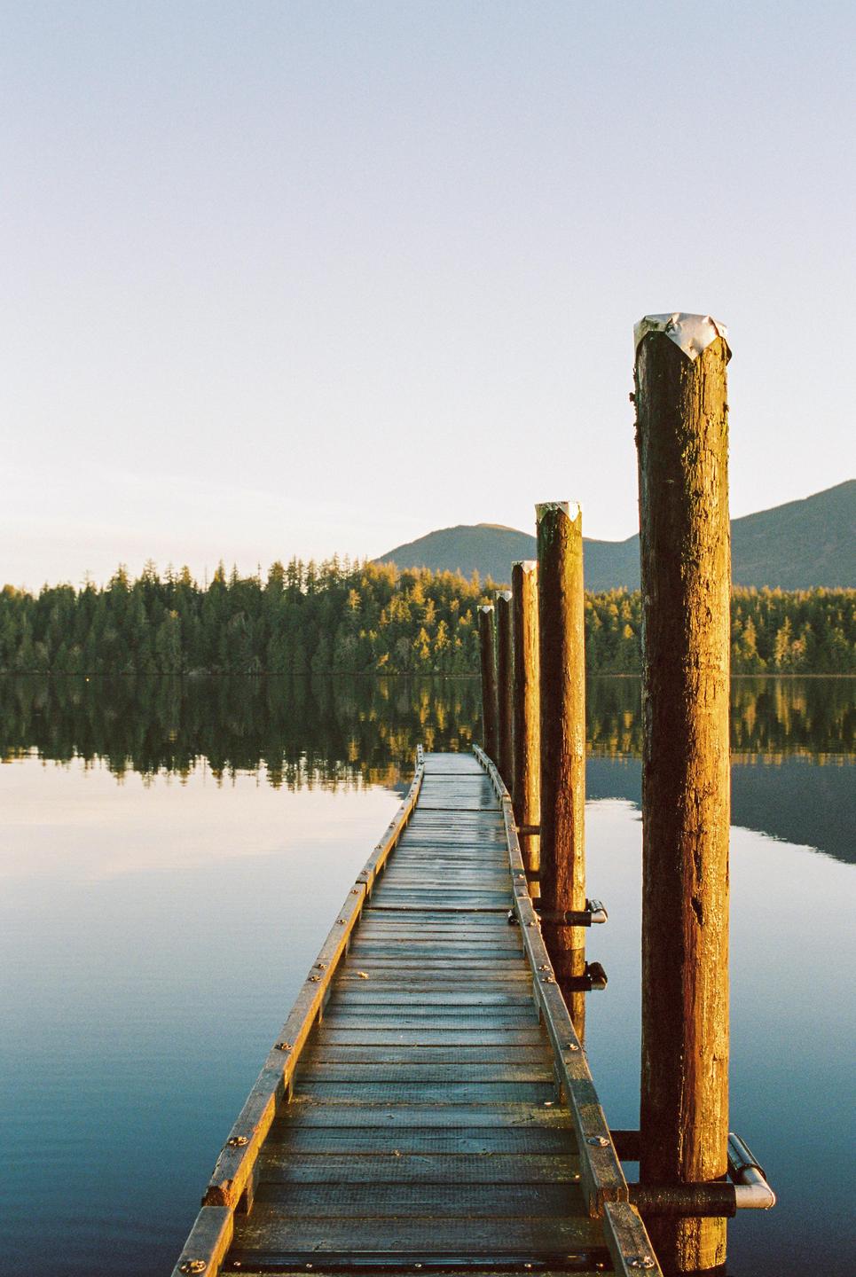 A wooden dock extends into a calm lake, reflecting the golden light of the setting sun on the surrounding forest and mountains.