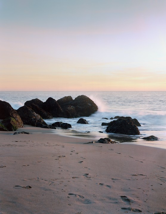 Waves crash against rocks on a sandy beach at sunset.