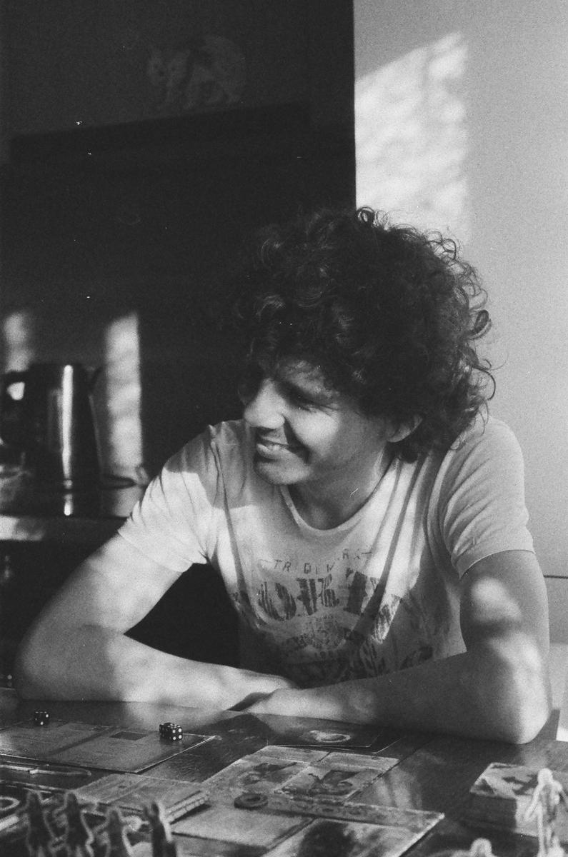 A man with curly hair sits at a table with board game pieces and dice, looking to the side and smiling.