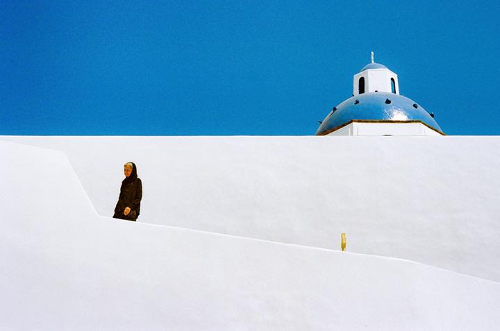 A woman in a dark coat stands on a white wall with a blue-domed building and clear blue sky in the background.
