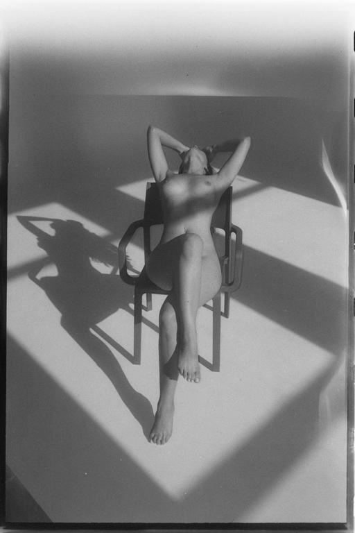 A nude woman reclines on a chair with her arms behind her head, casting a shadow on the wall and floor.