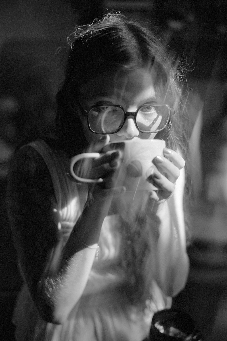 A woman wearing glasses sips from a mug, with light reflecting off her glasses and steam rising from the mug.