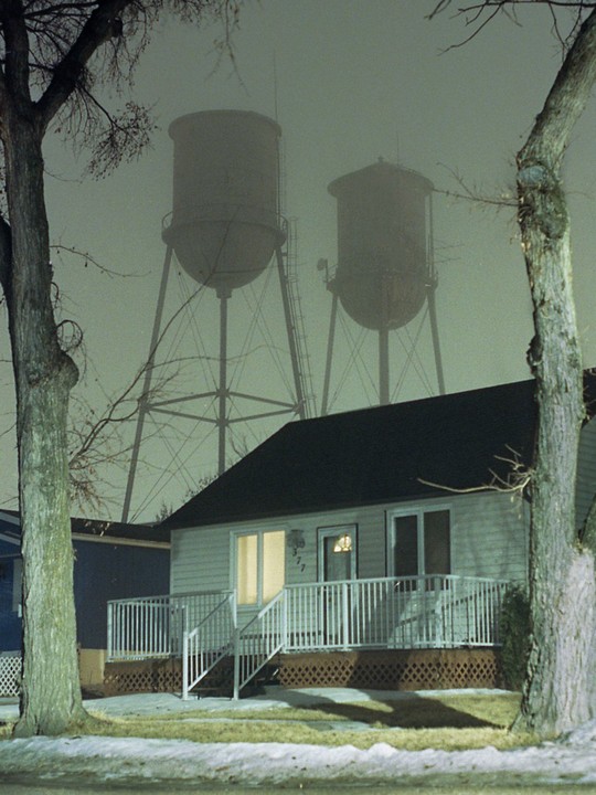 Two large water towers loom over a small house with a lit window at night.