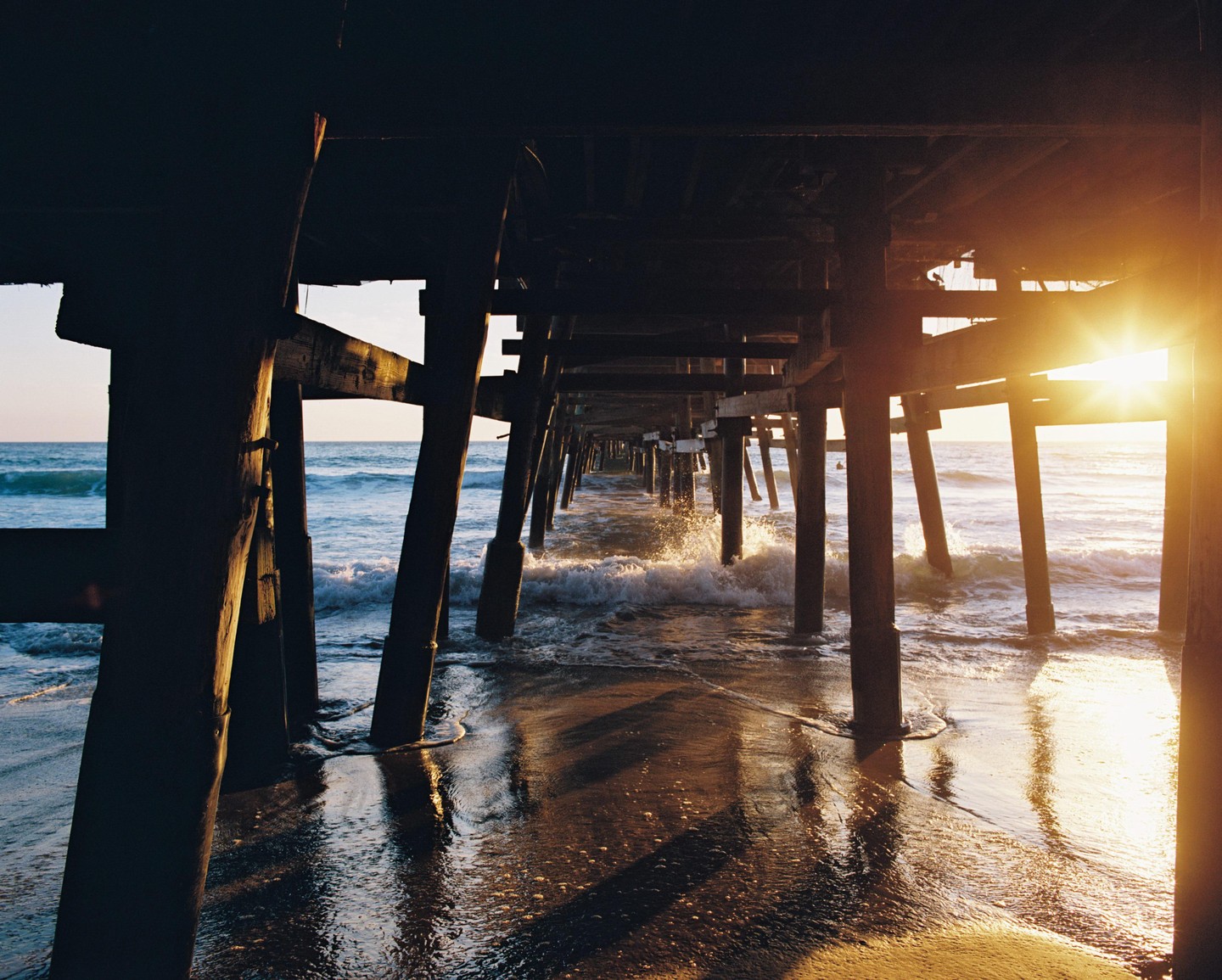 The sun shines brightly from behind a pier, illuminating the water and sand beneath it.