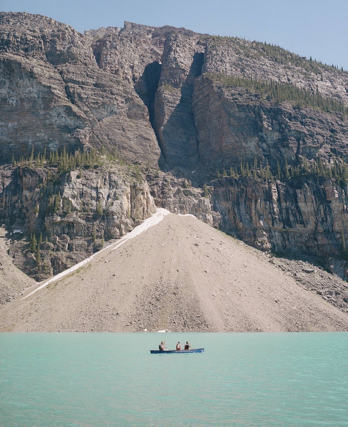 Three people in a canoe paddle across a turquoise lake in front of a large mountain.